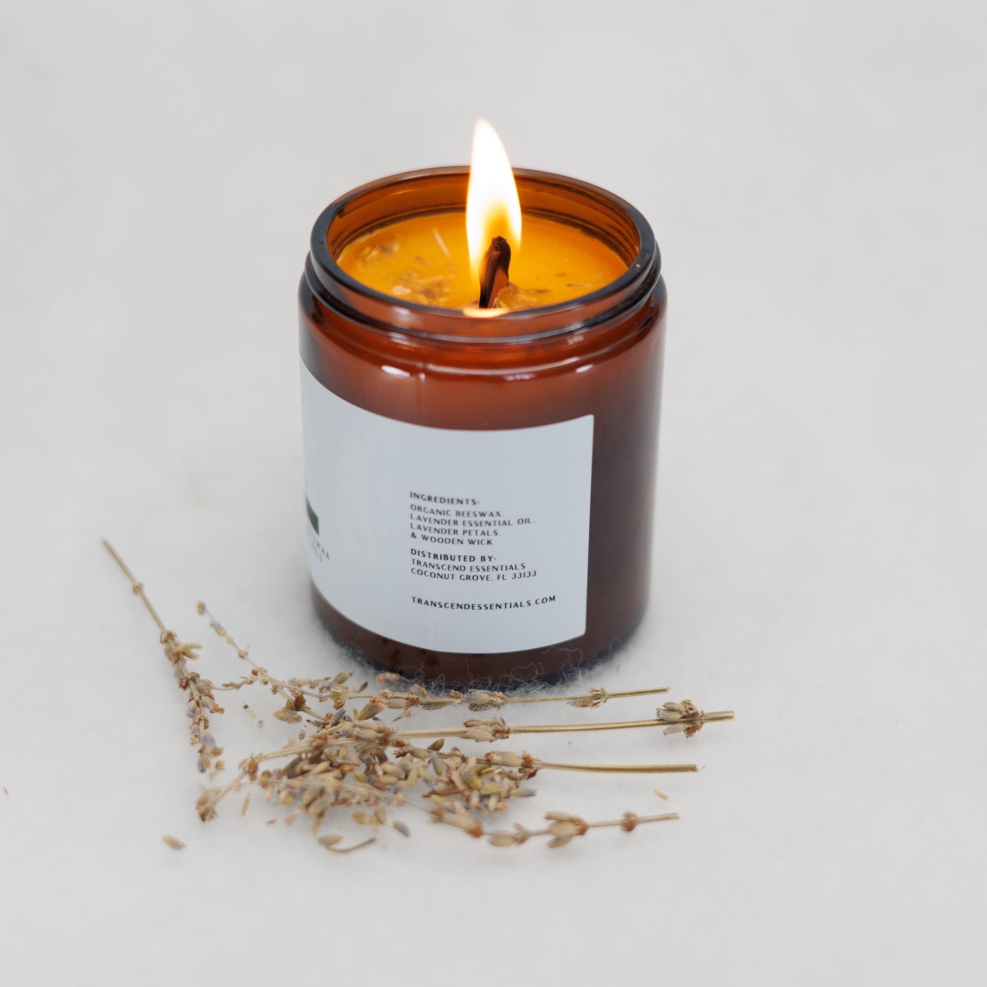 A lit amber glass candle showing the ingredients label sits on a light surface, with dried lavender sprigs placed in front of it. The label lists the ingredients as organic beeswax, lavender essential oil, lavender petals, and a wooden wick. The candle is made by Transcend Essentials.