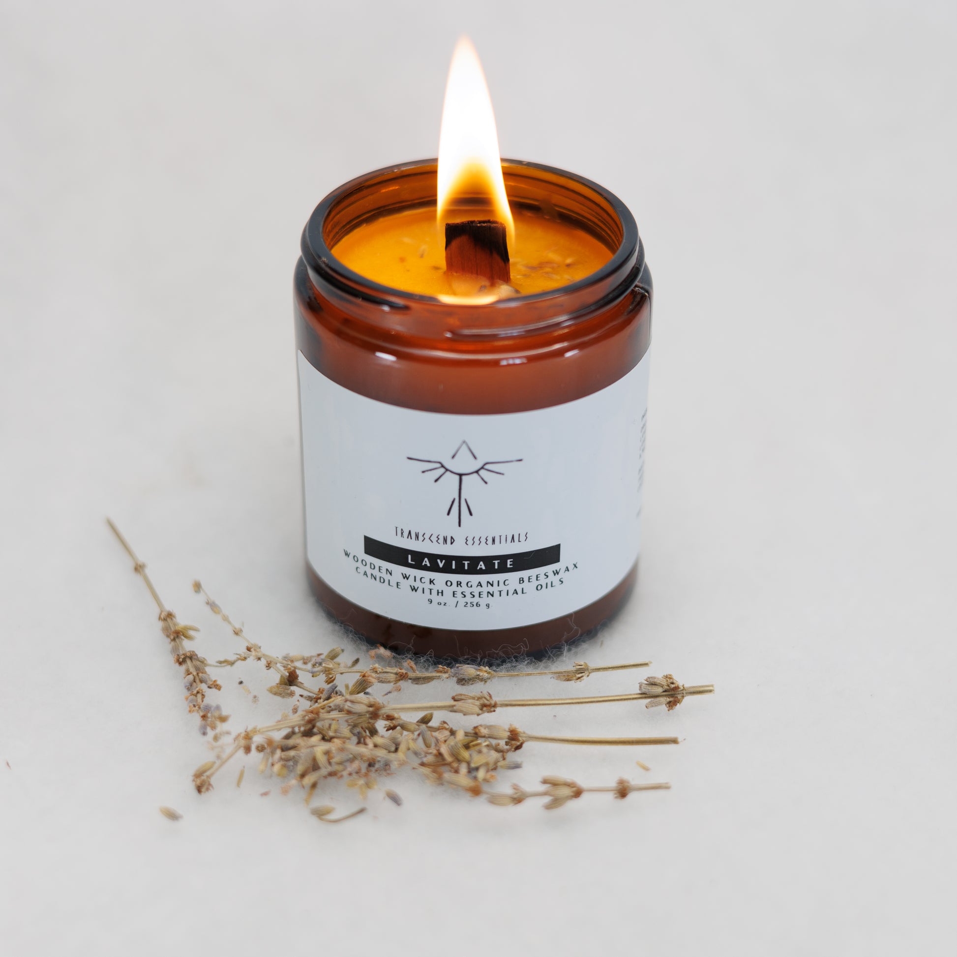 A lit amber glass candle labeled “Transcend Essentials – Lavitate” sits on a light surface, with dried lavender sprigs placed in front of it. The candle features a wooden wick and is made from organic beeswax with lavender essential oil.