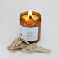 A lit amber glass candle sits on a light surface with a small pile of dried eucalyptus leaves in front of it. The label lists ingredients including organic beeswax, eucalyptus essential oil, eucalyptus leaves, and a wooden wick, along with distributor details for Transcend Essentials.