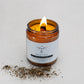 A lit amber glass candle labeled “Transcend Essentials – Peppermint” sits on a light surface, with a small pile of dried peppermint leaves in front of it. The candle features a wooden wick and is made from organic beeswax with Peppermint essential oil.