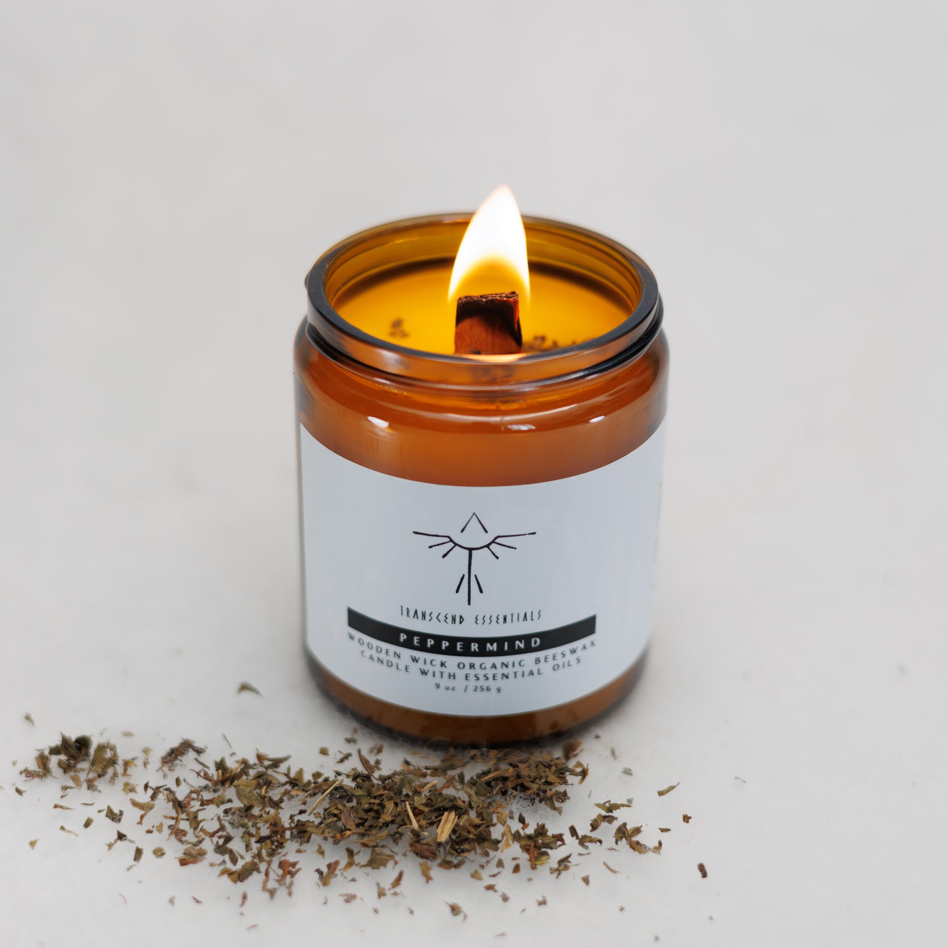 A lit amber glass candle labeled “Transcend Essentials – Peppermint” sits on a light surface, with a small pile of dried peppermint leaves in front of it. The candle features a wooden wick and is made from organic beeswax with Peppermint essential oil.